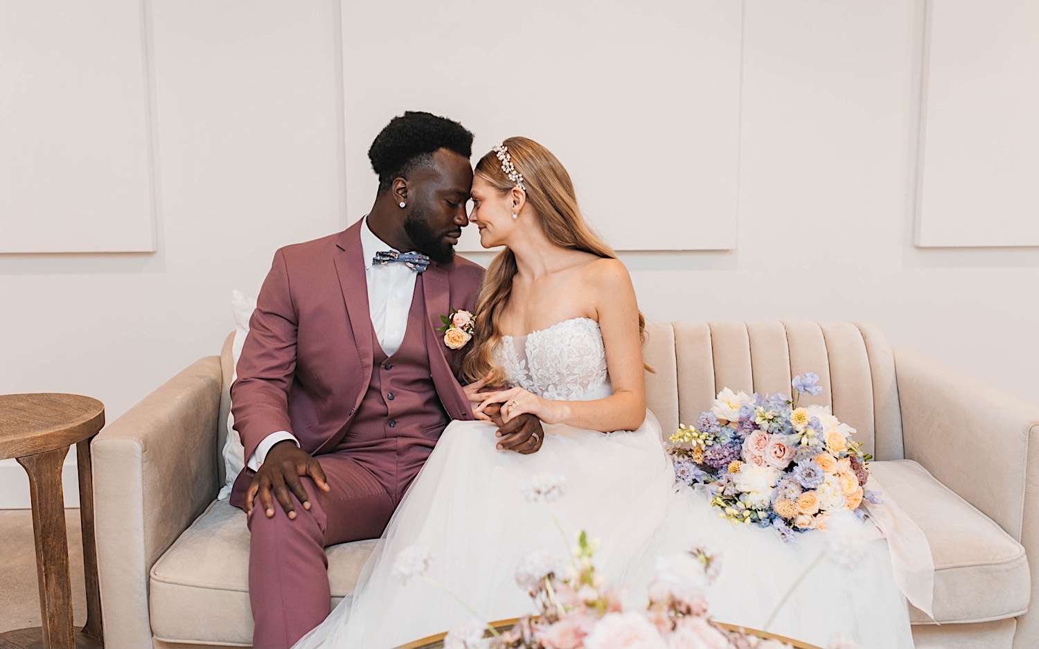 A bride and groom sit next to one another on a couch and touch their foreheads together during their wedding day at The Aisling