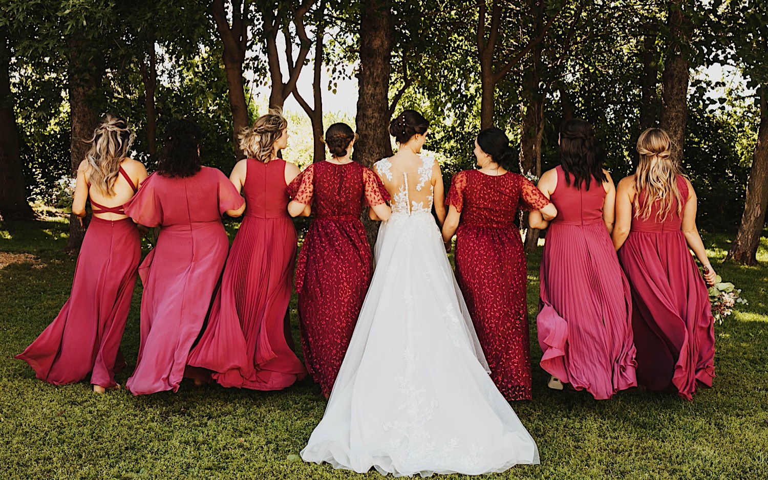 A bride and her bridesmaids lock arms and face away from the camera towards a row of trees after an outdoor wedding ceremony at Legacy Hill Farm