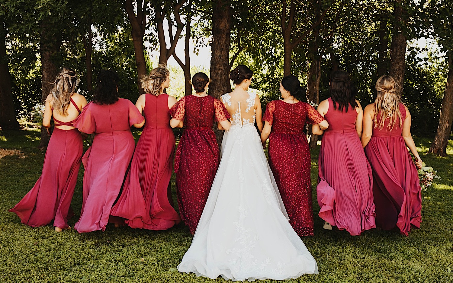 A bride and her bridesmaids lock arms and face away from the camera towards a row of trees after an outdoor wedding ceremony at Legacy Hill Farm A bride and her bridesmaids lock arms and face away from the camera towards a row of trees after an outdoor wedding ceremony at Legacy Hill Farm