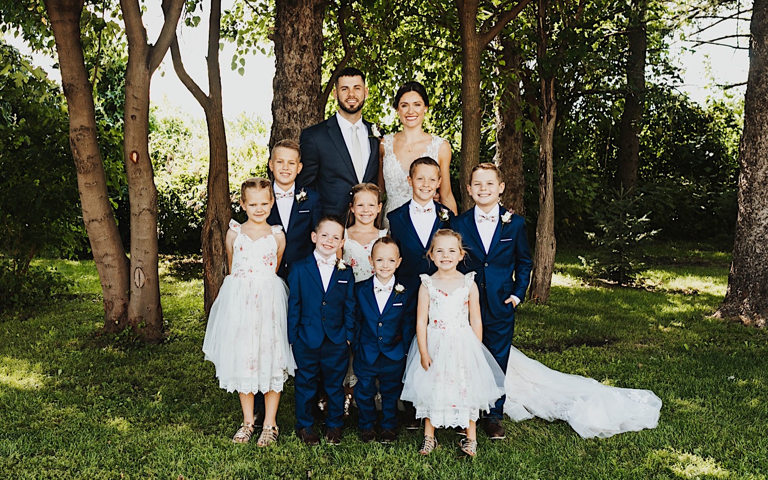 A bride and groom stand with a group of young children in front of a cluster of trees after their wedding ceremony at Legacy Hill Farm