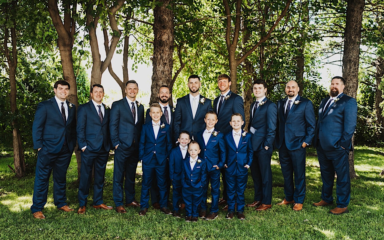 A groom stands with his groomsmen and the ring bearers as they all smile at the camera while in front of a row of trees after a wedding ceremony at Legacy Hill Farm