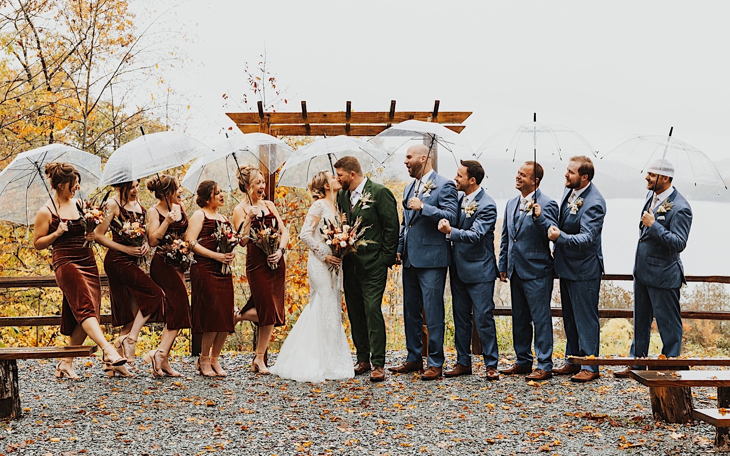 A bride and groom kiss while members of their wedding parties cheer for them on either side while they all stand at the outdoor ceremony space of Lake Bomoseen Lodge in Vermont