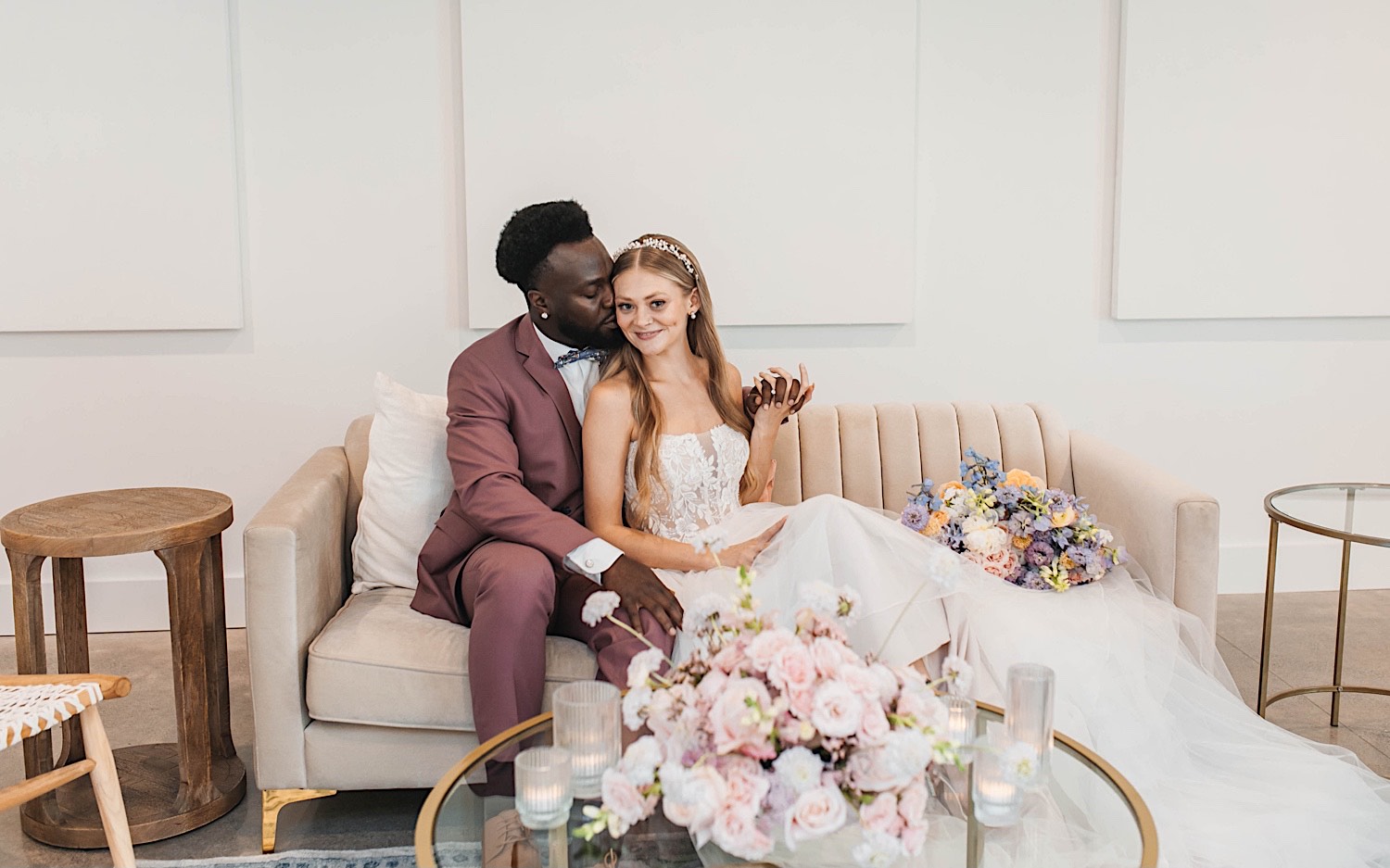 A bride smiles at the camera as the groom kisses her cheek while they sit on a couch during their wedding day at The Aisling