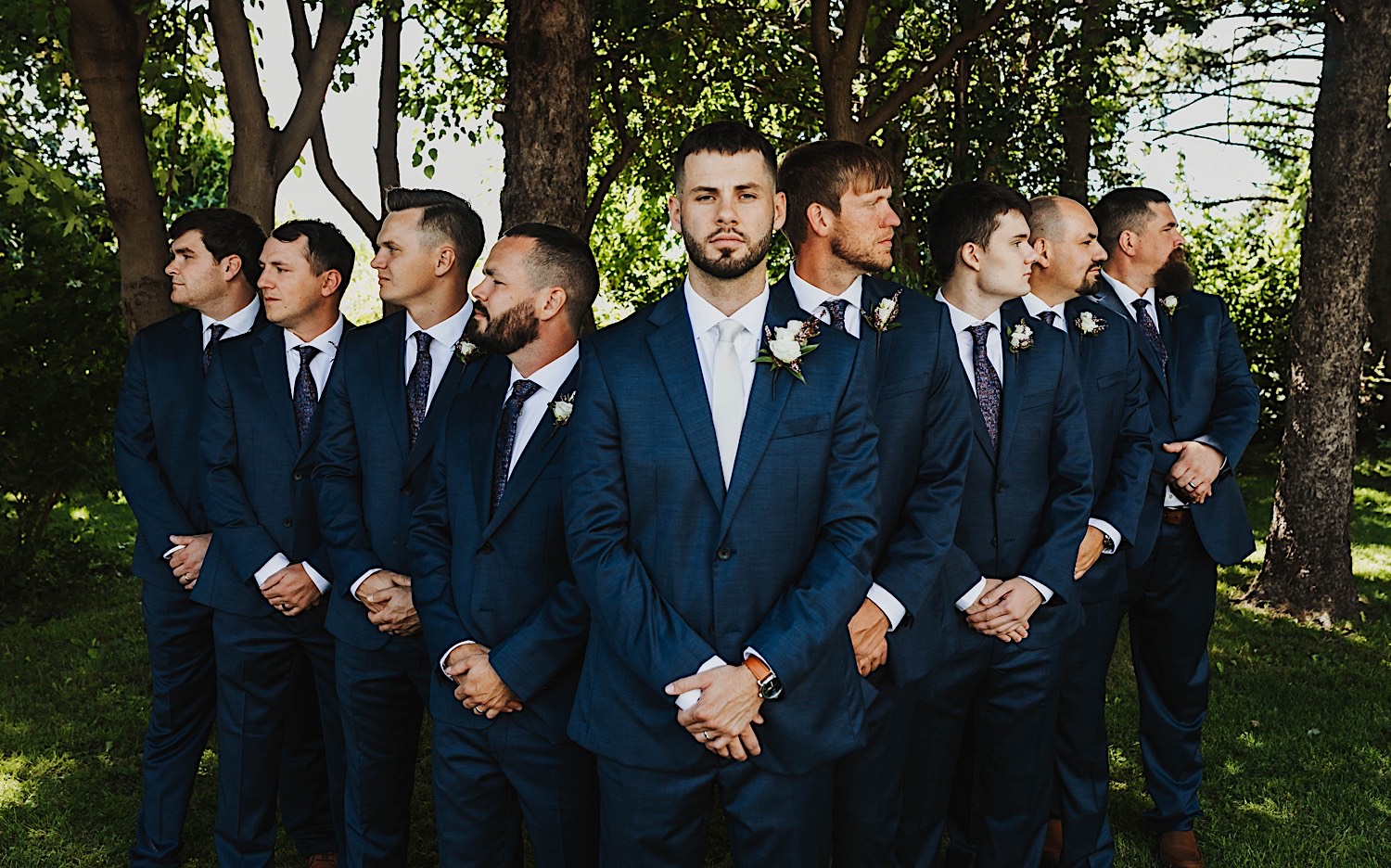 A groom faces the camera while his groomsmen on either side of him look away from the camera while standing in front of a row of trees at the wedding venue Legacy Hill Farm