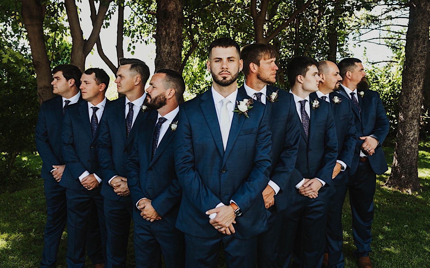 A groom faces the camera while his groomsmen on either side of him look away from the camera while standing in front of a row of trees at the wedding venue Legacy Hill Farm A groom faces the camera while his groomsmen on either side of him look away from the camera while standing in front of a row of trees at the wedding venue Legacy Hill Farm