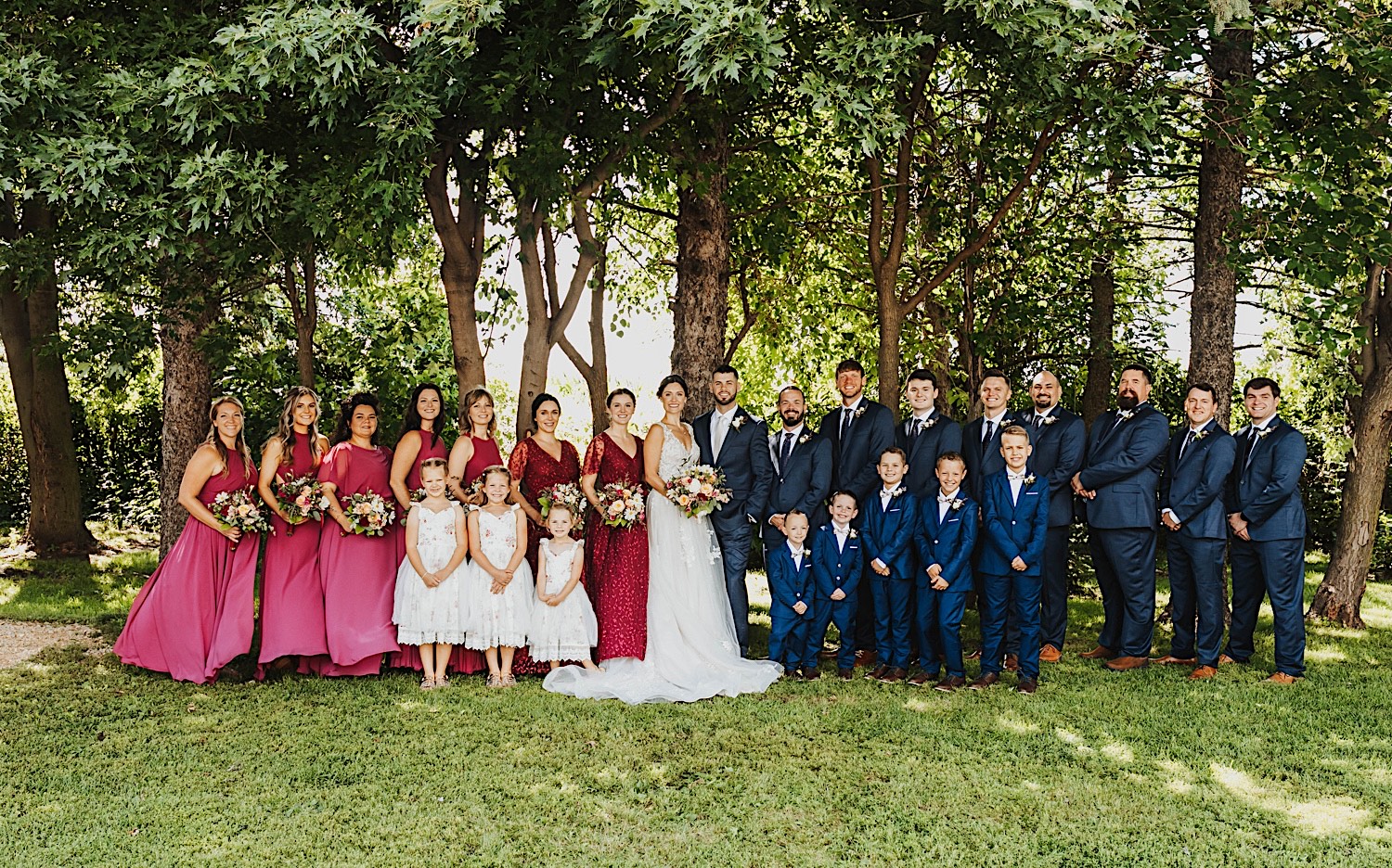 A bride and groom stand with their wedding parties, flower girls, and ring bearers in front of a row of trees at the wedding venue Legacy Hill Farm