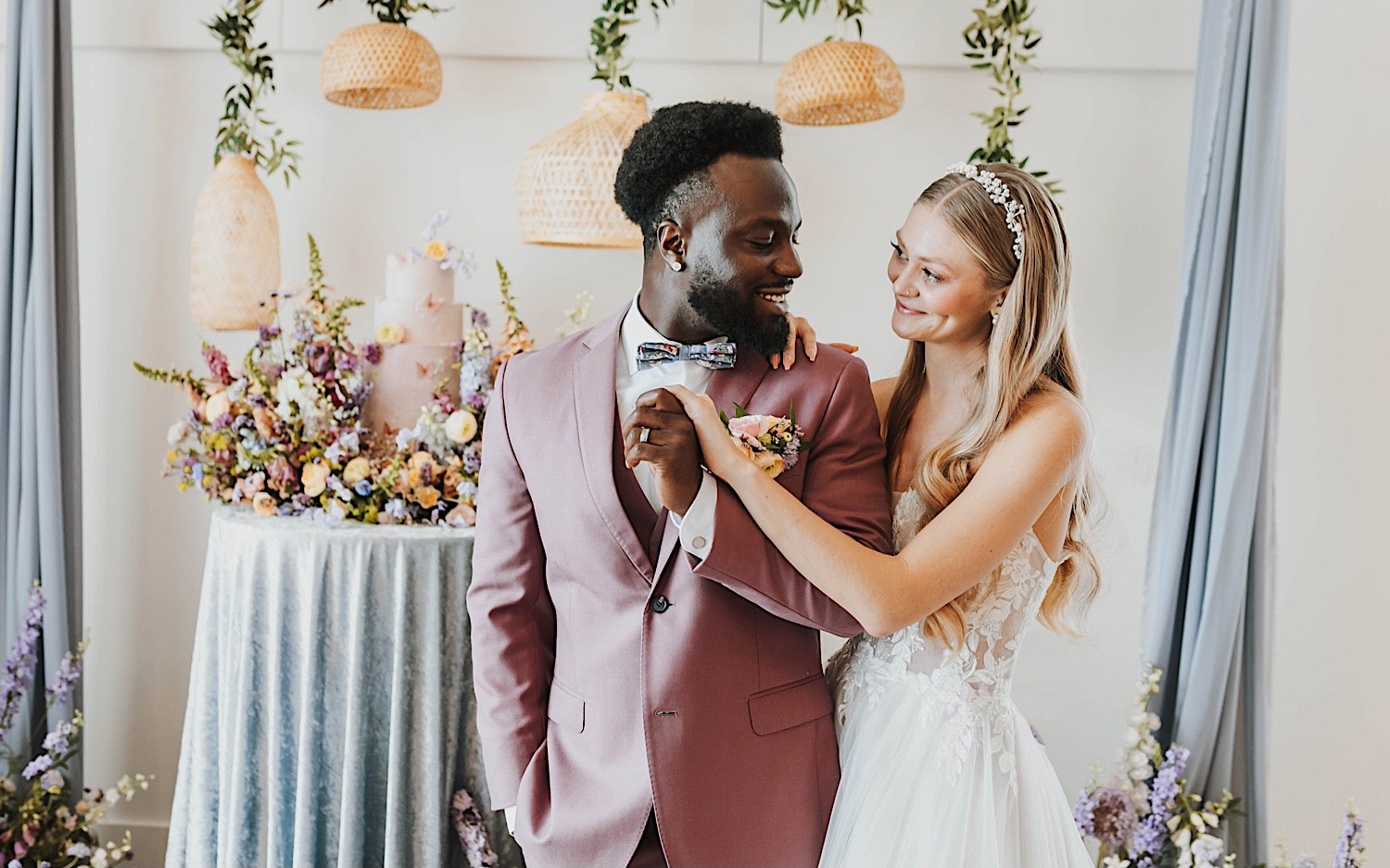 A bride and groom smile at one another while standing in front of their wedding cake during their wedding reception at The Aisling