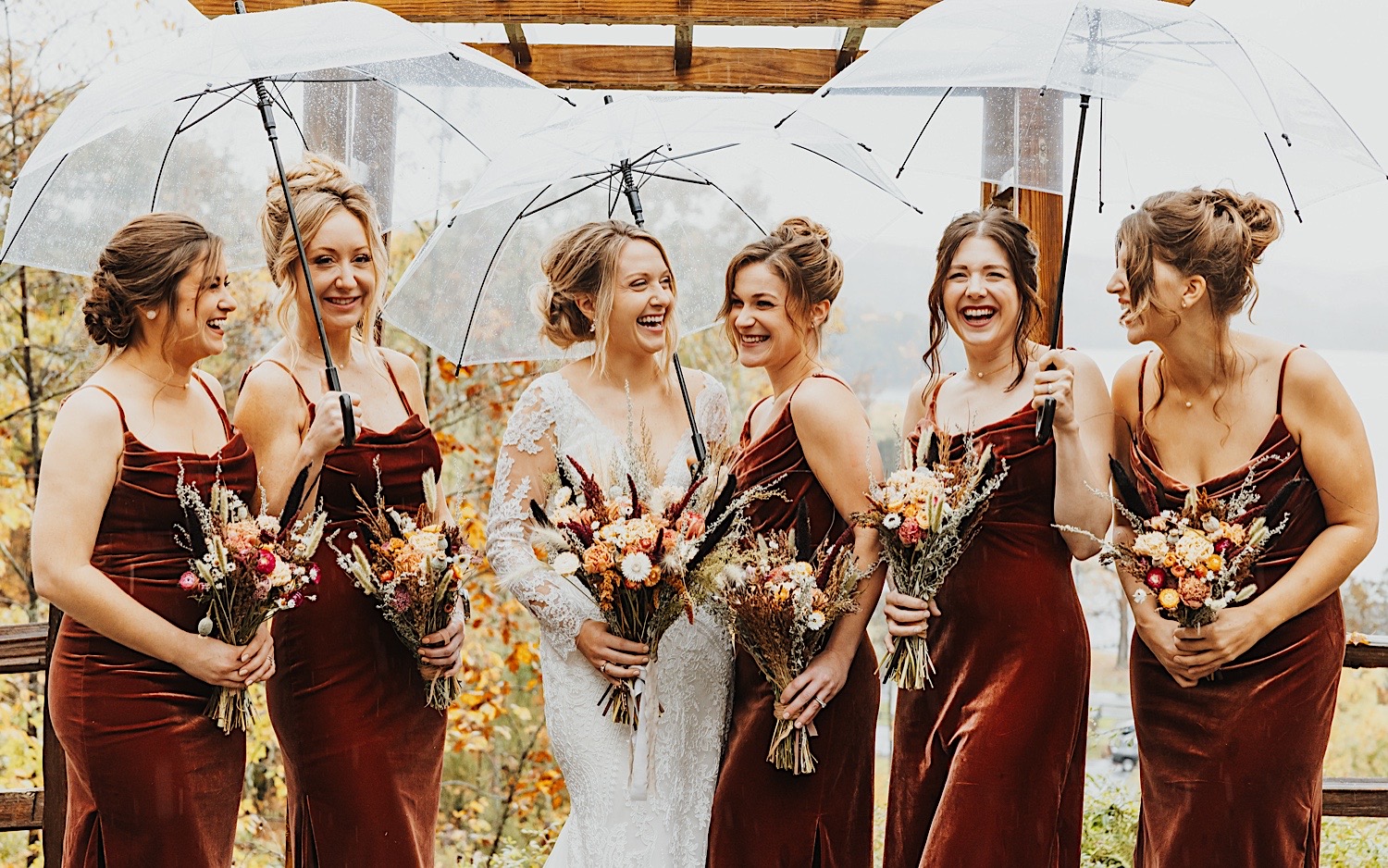 A bride laughs alongside her 5 bridesmaids while they take portrait photos in the rain at the outdoor wedding ceremony space of Lake Bomoseen Lodge in Vermont