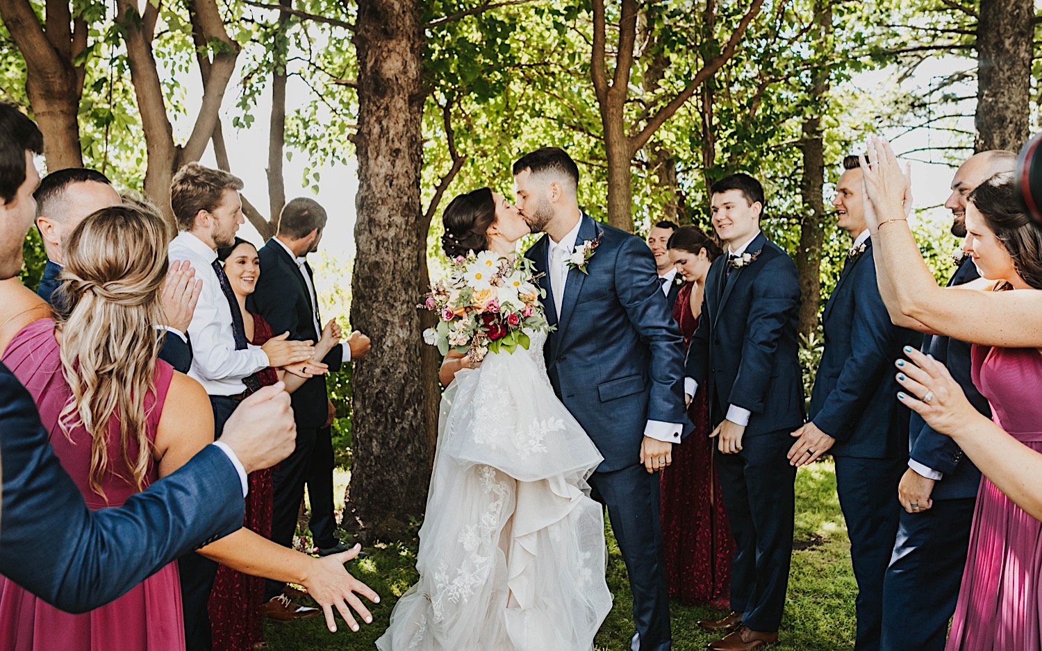 A bride and groom kiss while members of their wedding party surround them and cheer in front of a row of trees at the wedding venue Legacy Hill Farm