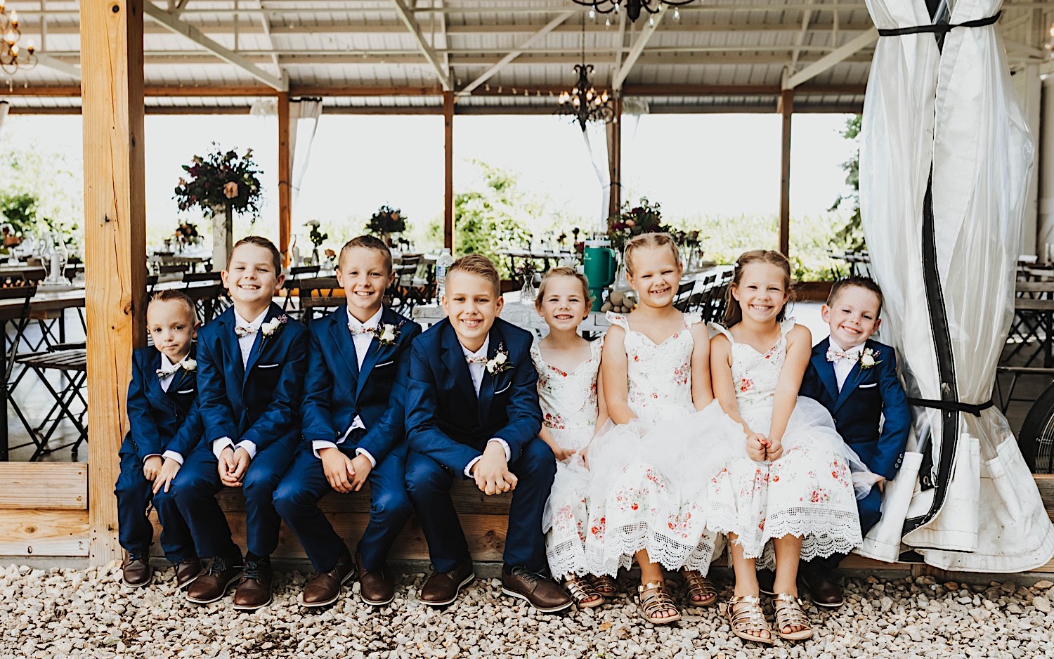 A group of children dressed for a wedding sit and smile at the camera while outside the reception space of the wedding venue Legacy Hill Farm