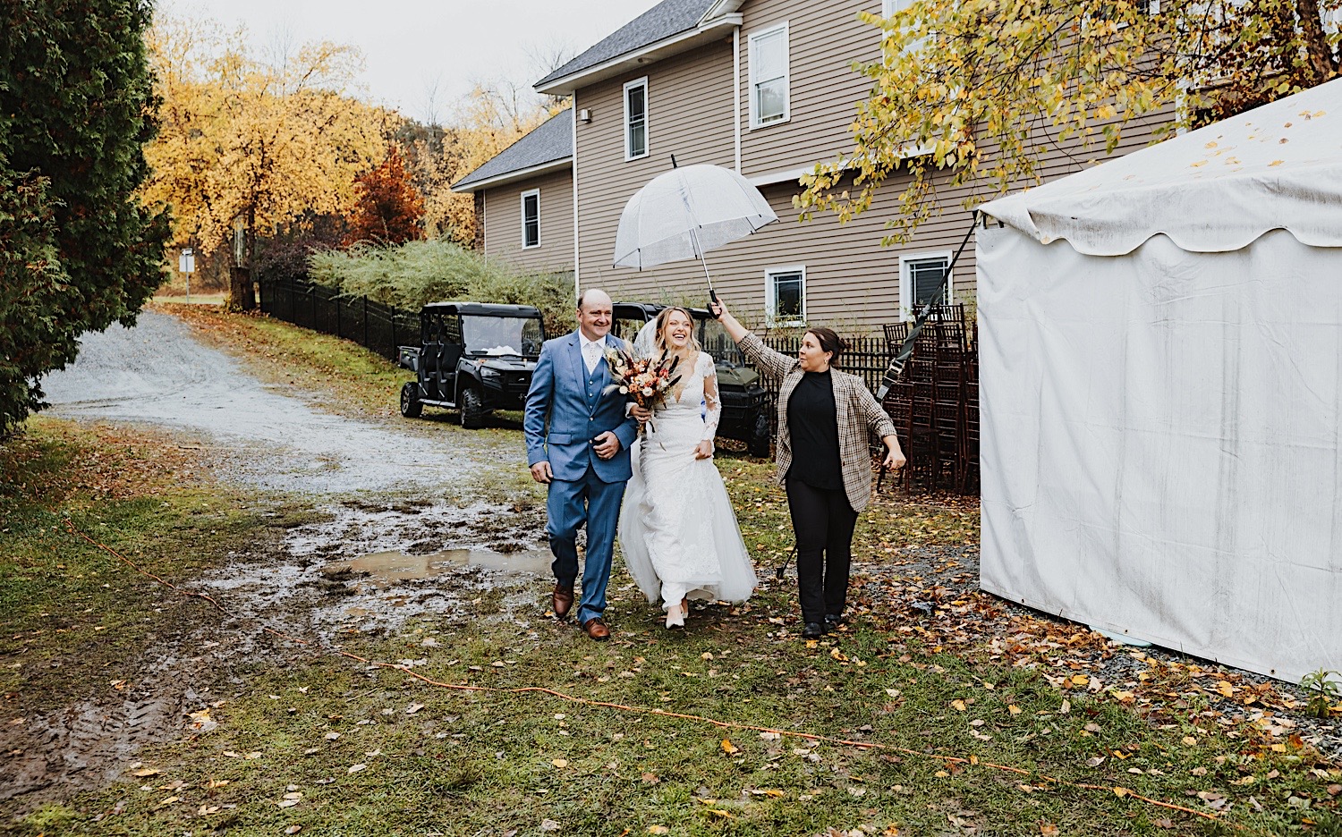 A bride laughs while walking towards the ceremony space for her wedding at Lake Bomoseen Lodge in Vermont, she is escorted by her father and a woman is holding an umbrella over the bride's head