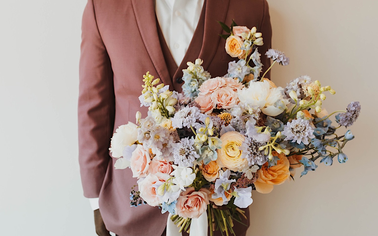 Torso photo of a groom standing and holding a flower bouquet towards the camera