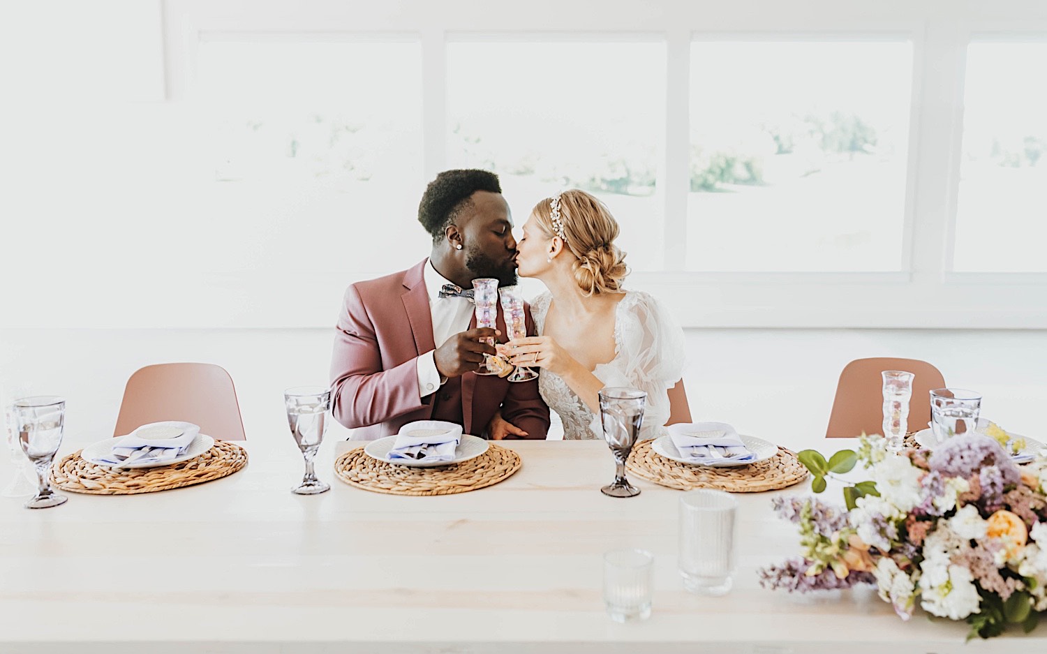 A bride and groom seated at their table kiss one another and clink their glasses together during their wedding reception at The Aisling