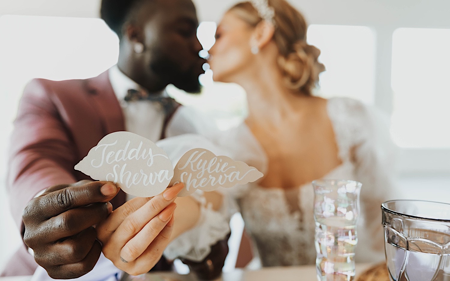 A bride and groom sit and kiss one another while holding their nametags towards the camera during their wedding reception at The Aisling