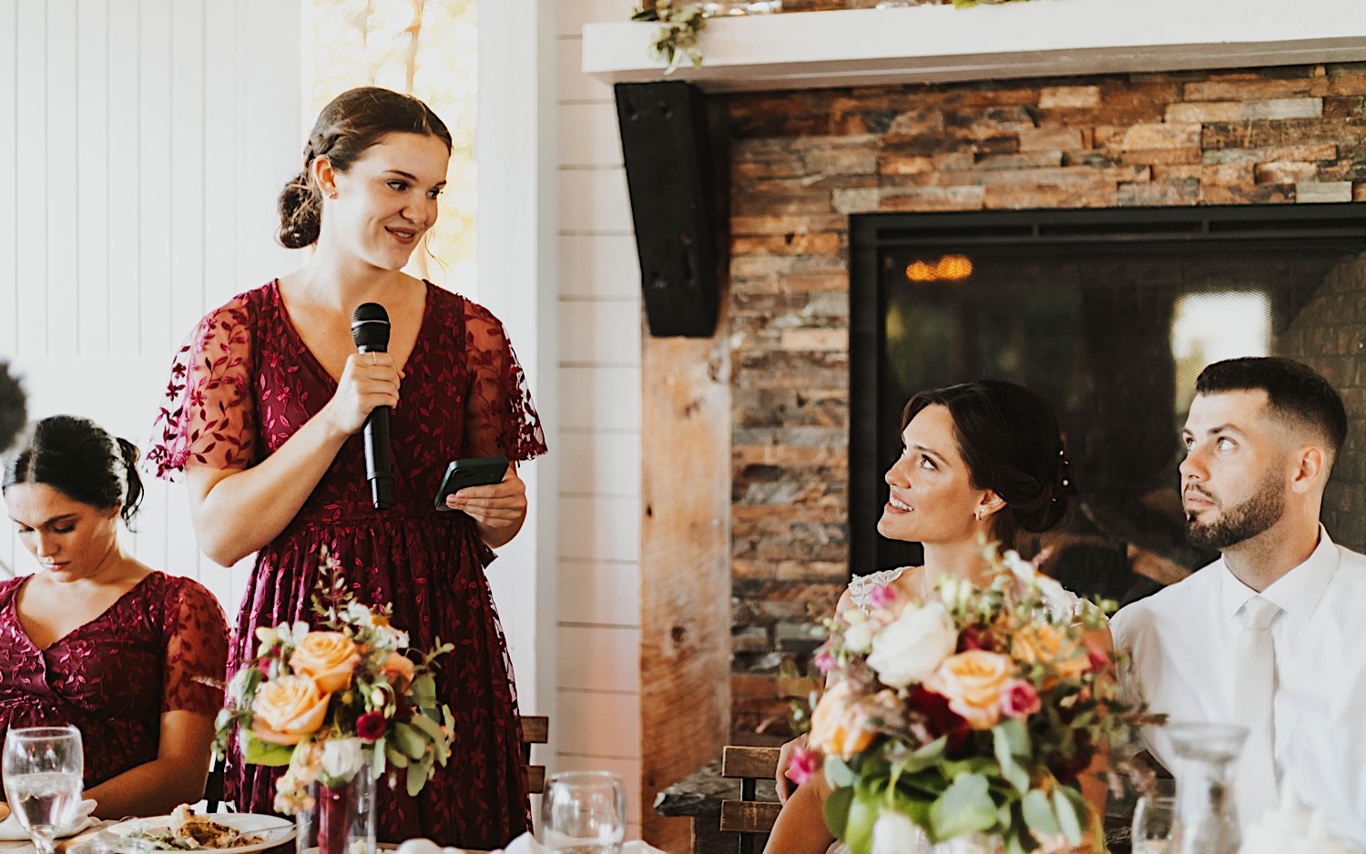 A bride and groom sit next to one another and look towards a standing bridesmaid as she gives a speech during their wedding reception at Legacy Hill Farm