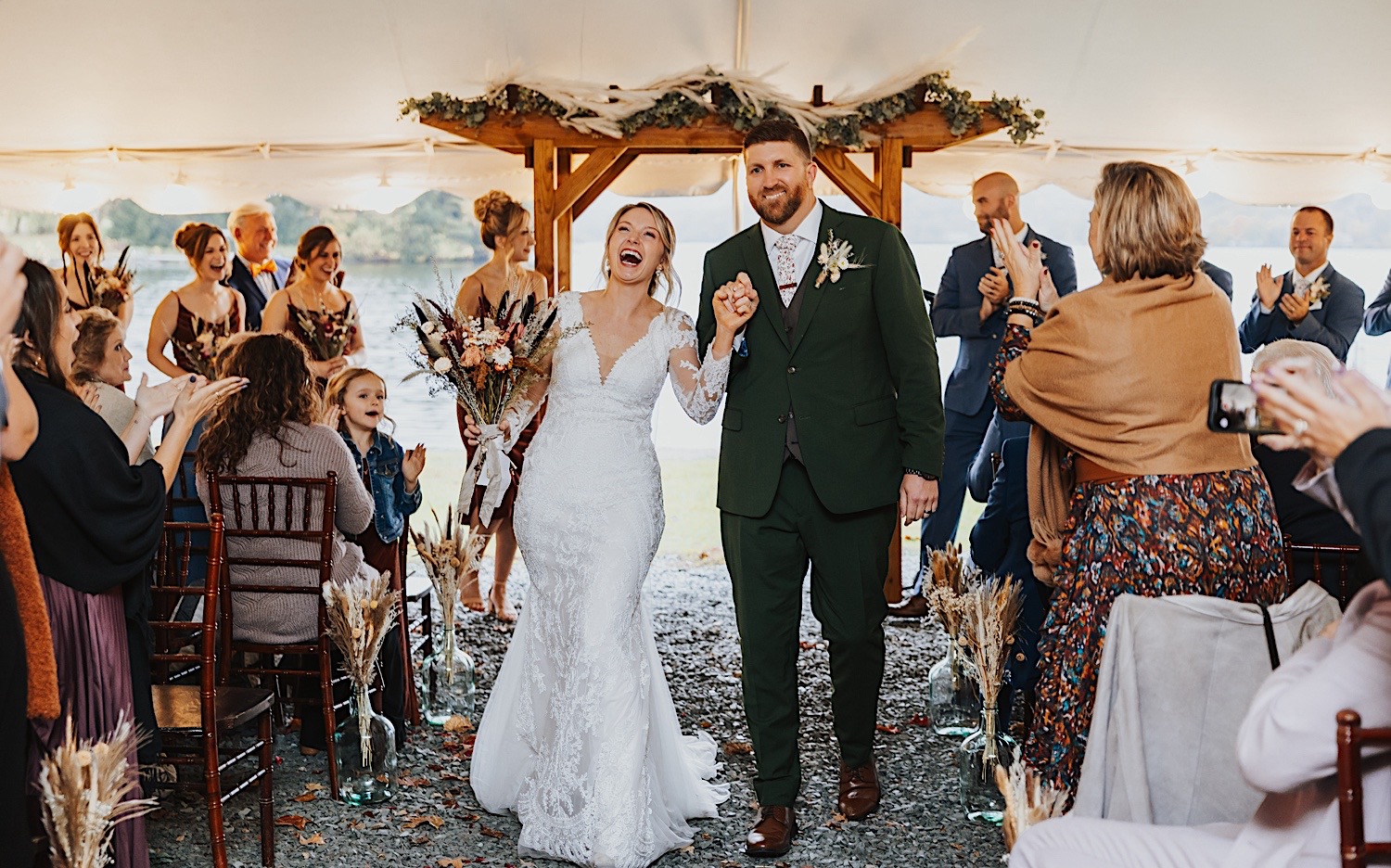 A bride and groom hold hands and smile while exiting their wedding ceremony under a tent on a rainy day at Lake Bomoseen Lodge in Vermont