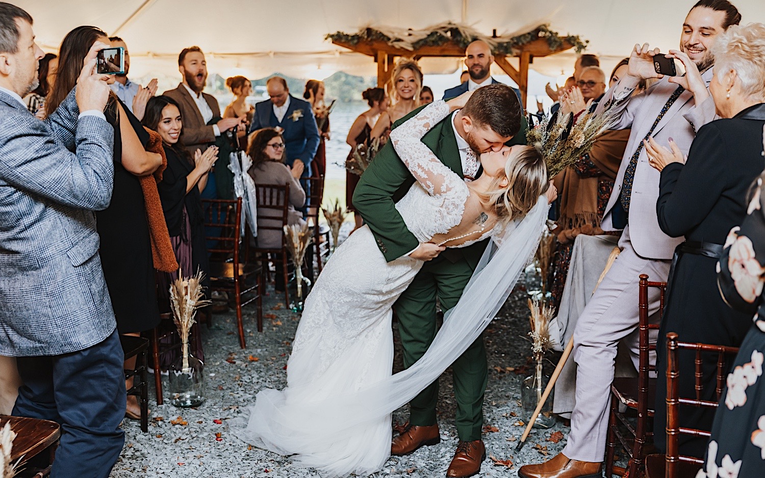 A bride and groom kiss while exiting their wedding ceremony as guests around them cheer, it is a rainy day so the ceremony took place under a tent at Lake Bomoseen Lodge in Vermont