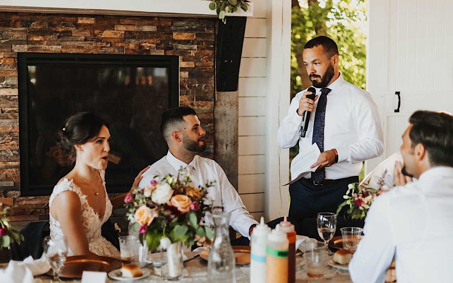 A bride and groom listen and sit next to one another while a groomsman gives a speech during their wedding reception at Legacy Hill Farm