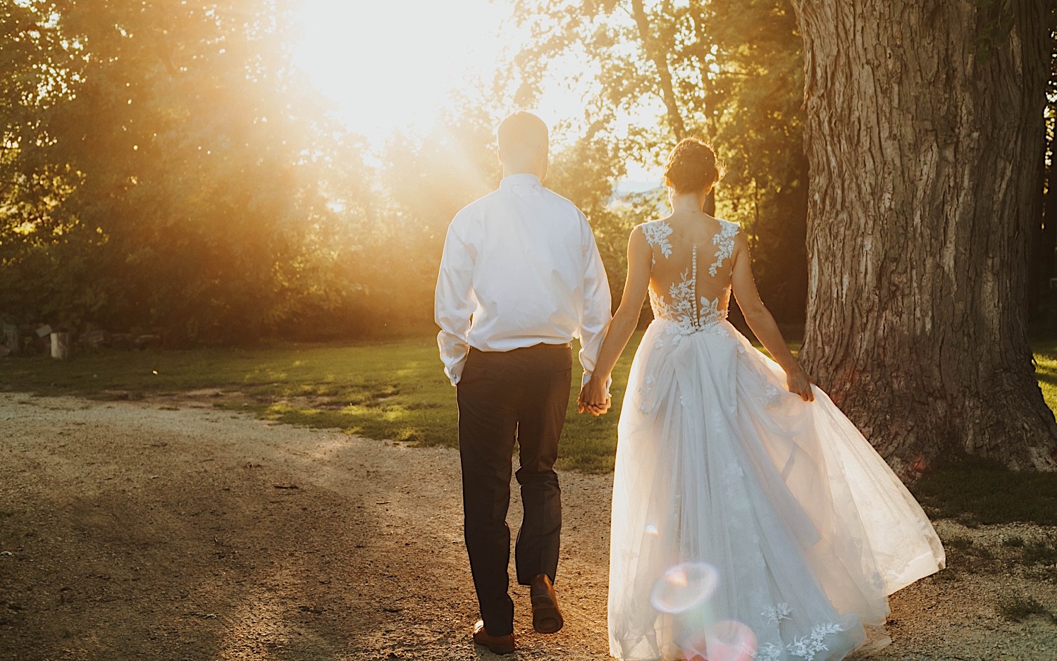 A bride and groom walk hand in hand along a dirt road away from the camera towards the sunset while at their wedding venue Legacy Hill Farm