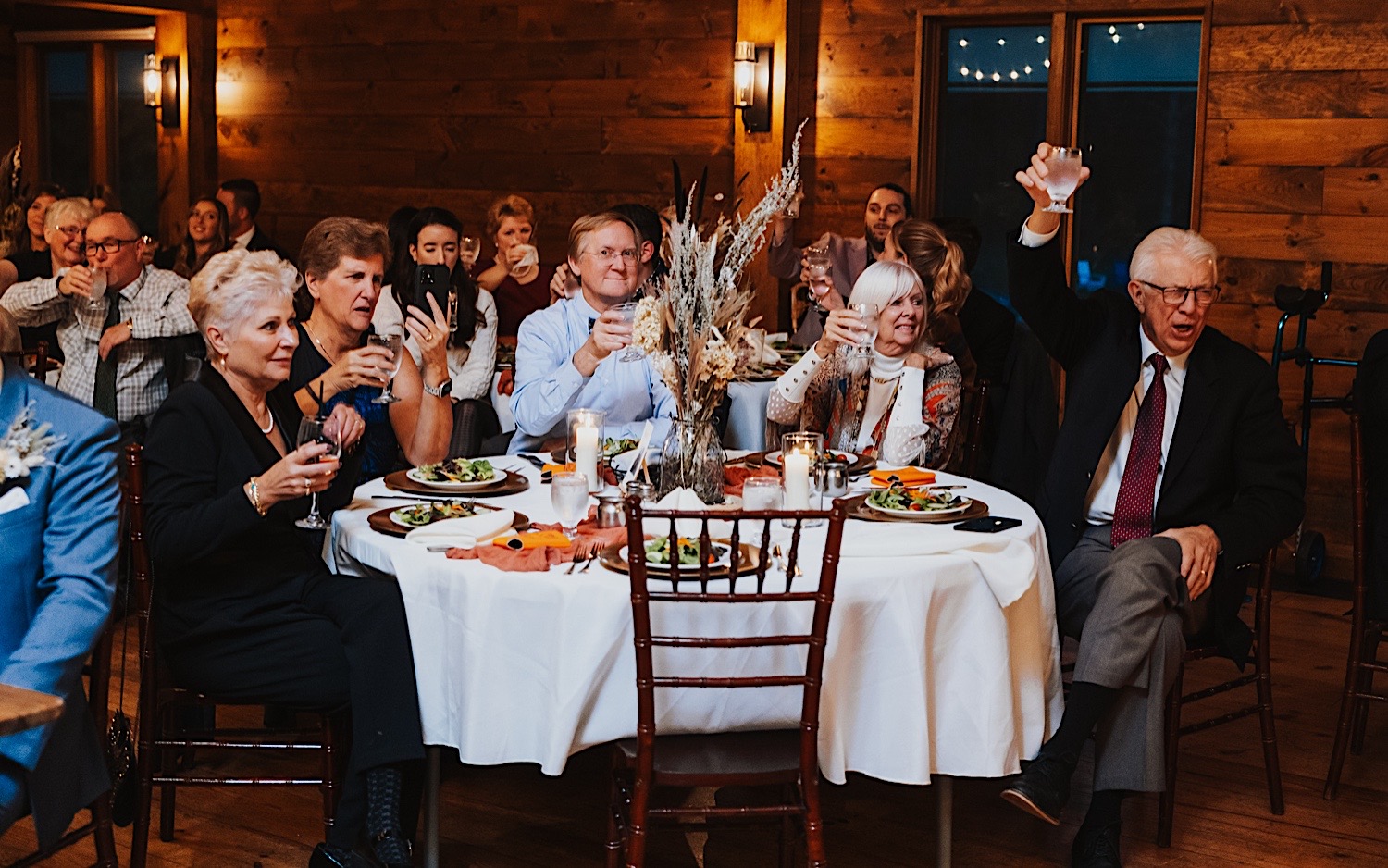 Guests of a wedding reception inside of Lake Bomoseen Lodge in Vermont raise their glasses and cheer while sitting at their table